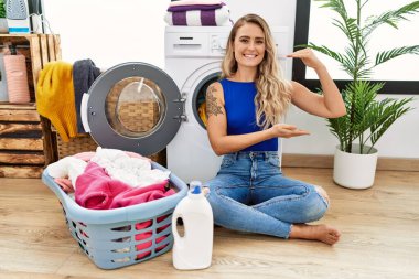 Young beautiful woman doing laundry sitting by wicker basket gesturing with hands showing big and large size sign, measure symbol. smiling looking at the camera. measuring concept. 