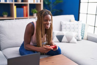 Evdeki kanepede oturan Afrikalı Amerikalı kadın video oyunu oynuyor.