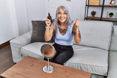 Orta yaşlı, kır saçlı, gece serumu kullanan bir kadın koltukta oturmuş gururla bağırıyor, zaferi kutluyor ve kollarını kaldırarak başarıyı kutluyor. 