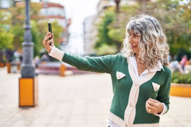 Orta yaşlı bir kadın, parktaki akıllı telefondan özçekim yaparken kendinden emin bir şekilde gülümsüyor.