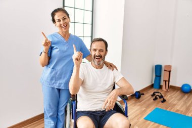 Hispanic middle age man sitting on wheelchair and nurse at rehabilitation clinic smiling happy pointing with hand and finger to the side 