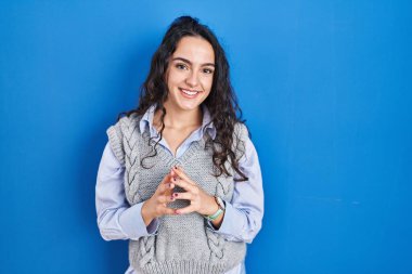 Young brunette woman standing over blue background hands together and fingers crossed smiling relaxed and cheerful. success and optimistic 