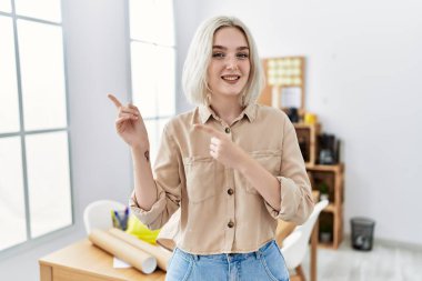 Young beautiful caucasian woman at construction office smiling and looking at the camera pointing with two hands and fingers to the side. 