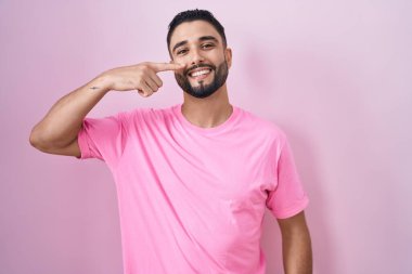 Pembe arka planda duran İspanyol genç adam, parmaklarıyla yüzünü ve burnunu gösteriyor, neşeli bir şekilde gülümsüyor. güzellik konsepti 