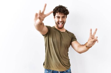 Hispanic man standing over isolated white background smiling with tongue out showing fingers of both hands doing victory sign. number two. 