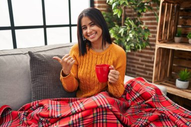 Genç İspanyol kadın kanepede oturup kahvesini yudumlarken arka tarafı işaret ediyor. El ve baş parmakları havada, kendinden emin gülümsüyor. 