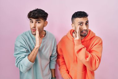 Genç İspanyol eşcinsel çift pembe arka planda duruyorlar ağızlarının üstünde gizli dedikodular söylüyorlar, kötü niyetli konuşmalar fısıldıyorlar. 