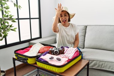 Beautiful middle age hispanic woman packing summer clothes in suitcase surprised with hand on head for mistake, remember error. forgot, bad memory concept. 