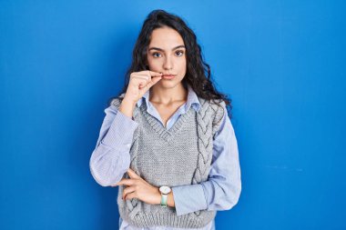 Young brunette woman standing over blue background mouth and lips shut as zip with fingers. secret and silent, taboo talking 