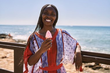 Genç Afrikalı Amerikalı kadın gülümseyerek sahilde dondurma yiyor..