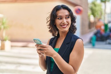 Genç, güzel, İspanyol bir kadın sokakta akıllı telefon kullanarak gülümsüyor.