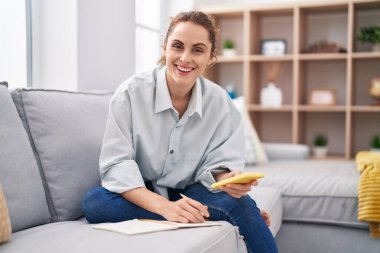 Young woman writing on notebook using smartphone at home