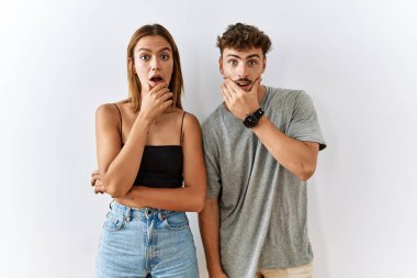 Young beautiful couple standing together over isolated background looking fascinated with disbelief, surprise and amazed expression with hands on chin 