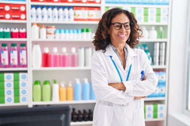 Middle age woman pharmacist smiling confident standing with arms crossed gesture at pharmacy