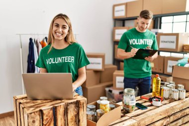 Young woman and man wearing volunteer t shirt at donations stand looking positive and happy standing and smiling with a confident smile showing teeth 