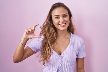 Pembe arka planda duran genç İspanyol kadın gülümsüyor ve el hareketleriyle el hareketleriyle küçük boy işareti yapıyor ve kameraya bakıyor. ölçüm kavramı. 