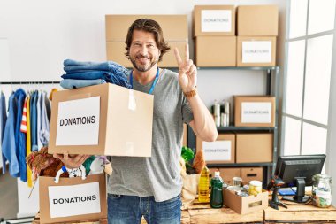 Handsome middle age man holding donations box for charity at volunteer stand smiling looking to the camera showing fingers doing victory sign. number two. 