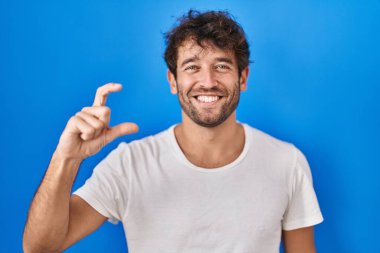 Hispanic young man standing over blue background smiling and confident gesturing with hand doing small size sign with fingers looking and the camera. measure concept. 