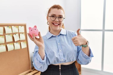 Ofiste domuz kumbarasını tutan genç kızıl saçlı kadın mutlu ve pozitif gülümsüyor, başparmağıyla mükemmel ve onay işareti yapıyor. 
