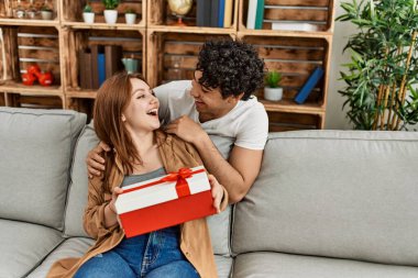 Mutlu gülümseyen genç çift evde yıldönümü hediyesiyle koltukta oturuyor..