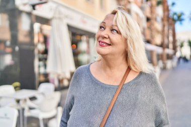 Orta yaşlı sarışın kadın sokakta kendinden emin bir şekilde gülümsüyor.