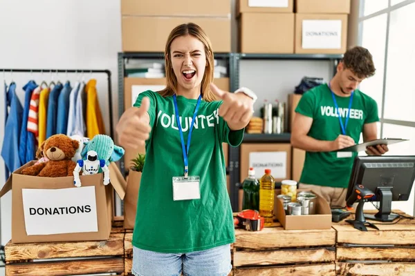 Young blonde girl wearing volunteer t shirt at donation stand approving doing positive gesture with hand, thumbs up smiling and happy for success. winner gesture.