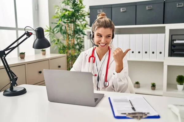 穿着医生制服的年轻医生女士在工作时 用电脑笔记本电脑把大拇指指向旁边 微笑着张开了嘴 — 图库照片