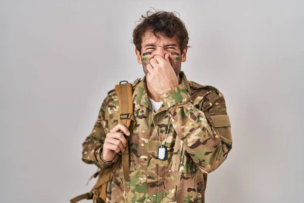 Hispanic young man wearing camouflage army uniform smelling something stinky and disgusting, intolerable smell, holding breath with fingers on nose. bad smell