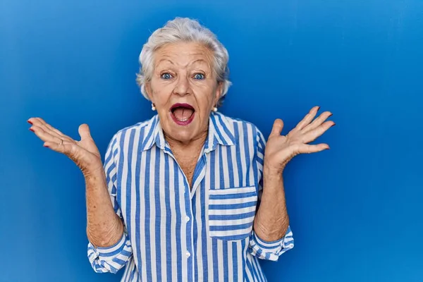 一位白发苍苍的老妇人站在蓝色的背景上 兴奋地欢呼着 欢呼着成功地举起双臂 睁开眼睛 获奖者概念 — 图库照片
