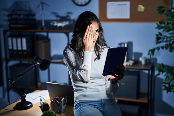 Young brazilian woman using touchpad at night working at the office yawning tired covering half face, eye and mouth with hand. face hurts in pain.