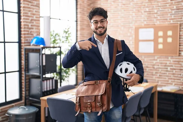 Hispanic man with beard holding bike helmet at the office pointing finger to one self smiling happy and proud
