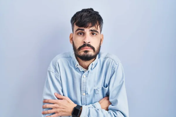 Young hispanic man with beard standing over blue background shaking and freezing for winter cold with sad and shock expression on face