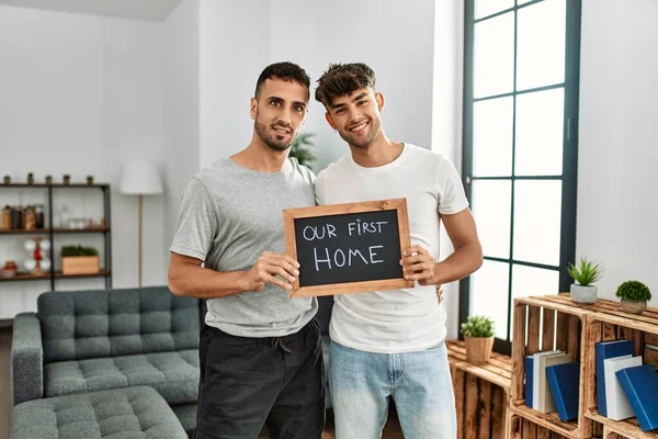 2人のヒスパニック系の男性カップル抱擁お互いに保持黒板で私たちの最初のホームメッセージで自宅 — ストック写真