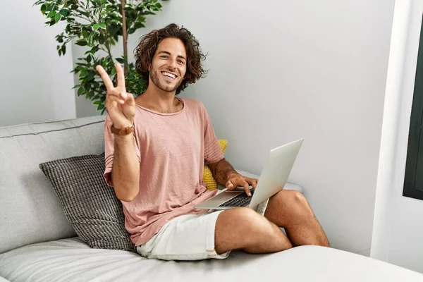 自宅のソファに腰掛けていた若いヒスパニック系の男は カメラを手で勝利のサインをする幸せな顔をして笑顔でノートパソコンを使用している — ストック写真