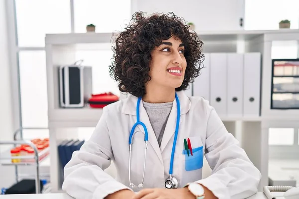 Young brunette woman with curly hair wearing doctor uniform and stethoscope looking to side, relax profile pose with natural face and confident smile.