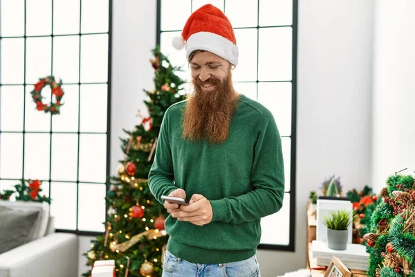 Young redhead man using smartphone standing by christmas tree at home