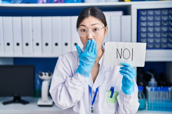Chinese young woman working at scientist laboratory holding no banner covering mouth with hand, shocked and afraid for mistake. surprised expression