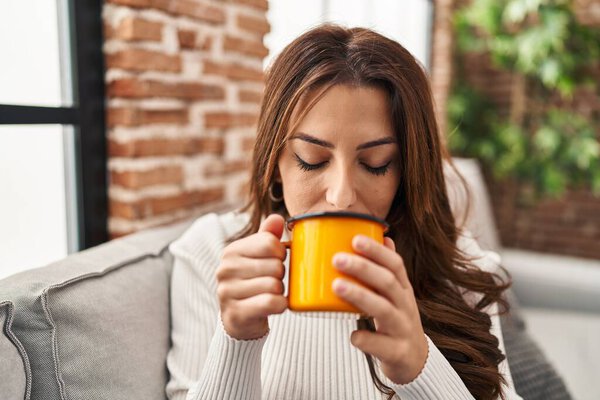 Young hispanic woman drinking coffee sitting on sofa at home