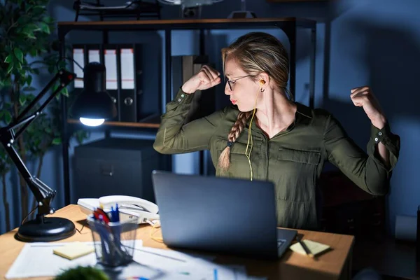 Young blonde woman working at the office at night showing arms muscles smiling proud. fitness concept.