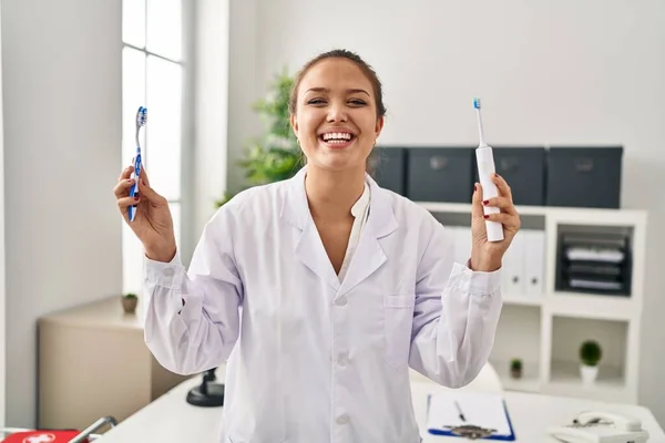 Young hispanic dentist woman holding ordinary toothbrush and electric toothbrush smiling and laughing hard out loud because funny crazy joke.