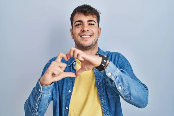 青い背景の上に立つ若いヒスパニック系の男が手でハートのシンボル形状を行う愛に笑みを浮かべて ロマンチックなコンセプト — ストック写真