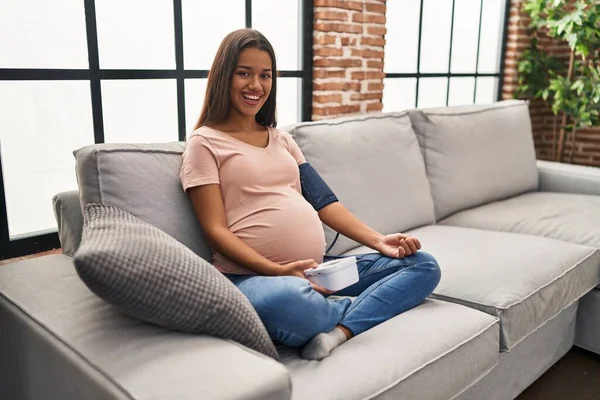 Młoda Kobieta Ciąży Pomocą Ciśnieniomierza Siedzi Kanapie Uśmiechając Się Śmiejąc — Zdjęcie stockowe