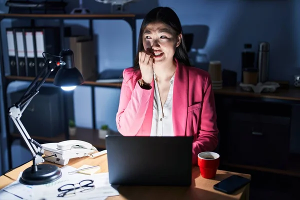 Chinese young woman working at the office at night looking stressed and nervous with hands on mouth biting nails. anxiety problem.