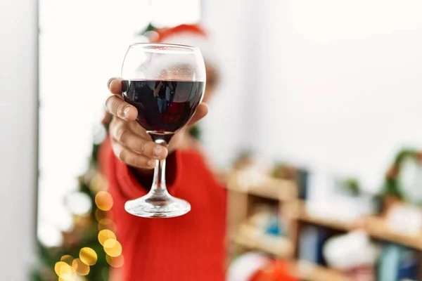 Young hispanic woman toasting with wine standing by christmas tree at home