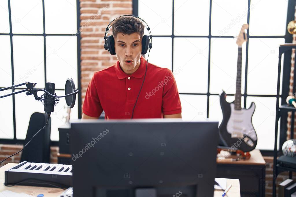 Young hispanic man playing piano keyboard at music studio scared and amazed with open mouth for surprise, disbelief face 