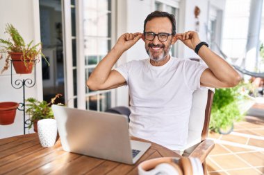 Middle age man using computer laptop at home smiling pulling ears with fingers, funny gesture. audition problem 