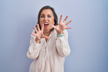 Middle age hispanic woman standing over blue background afraid and terrified with fear expression stop gesture with hands, shouting in shock. panic concept. 