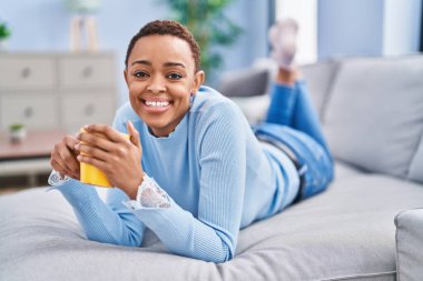 Afro-Amerikalı kadın evdeki kanepede kahve içiyor.