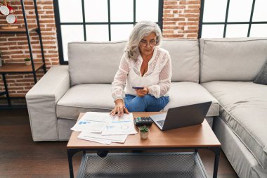 Evde dizüstü bilgisayar ve kredi kartı kullanan orta yaşlı kadın.