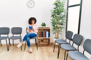 Orta Doğulu genç bir kadın bekleme odasında sandalyeye oturmuş dokunmatik ped kullanıyor.